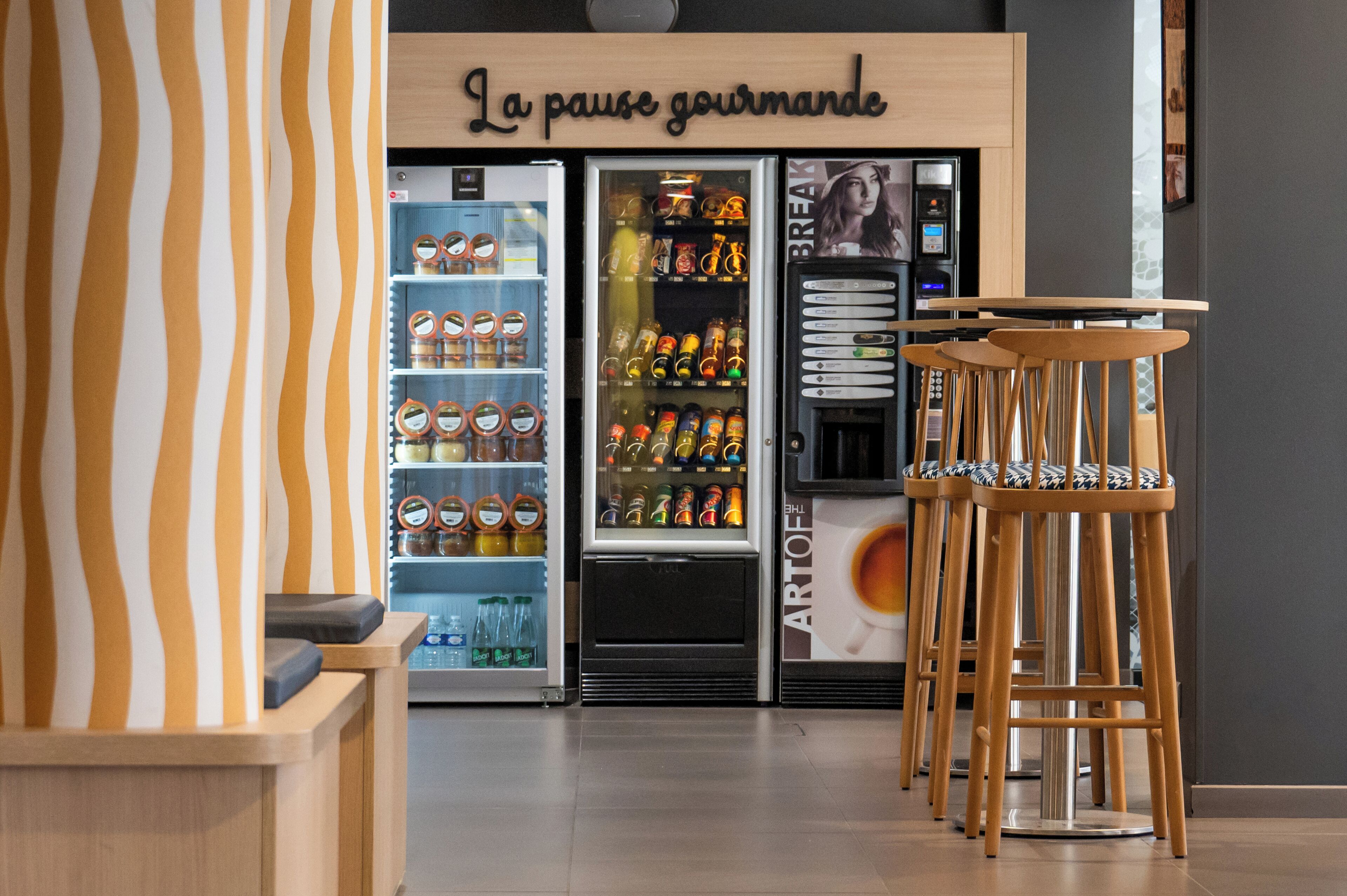 A café with a striped wall, a refrigerated display case with beverages, and wooden bar stools.