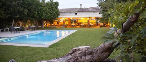 A swimming pool with a grassy area, outdoor seating, and a building with warm lighting.