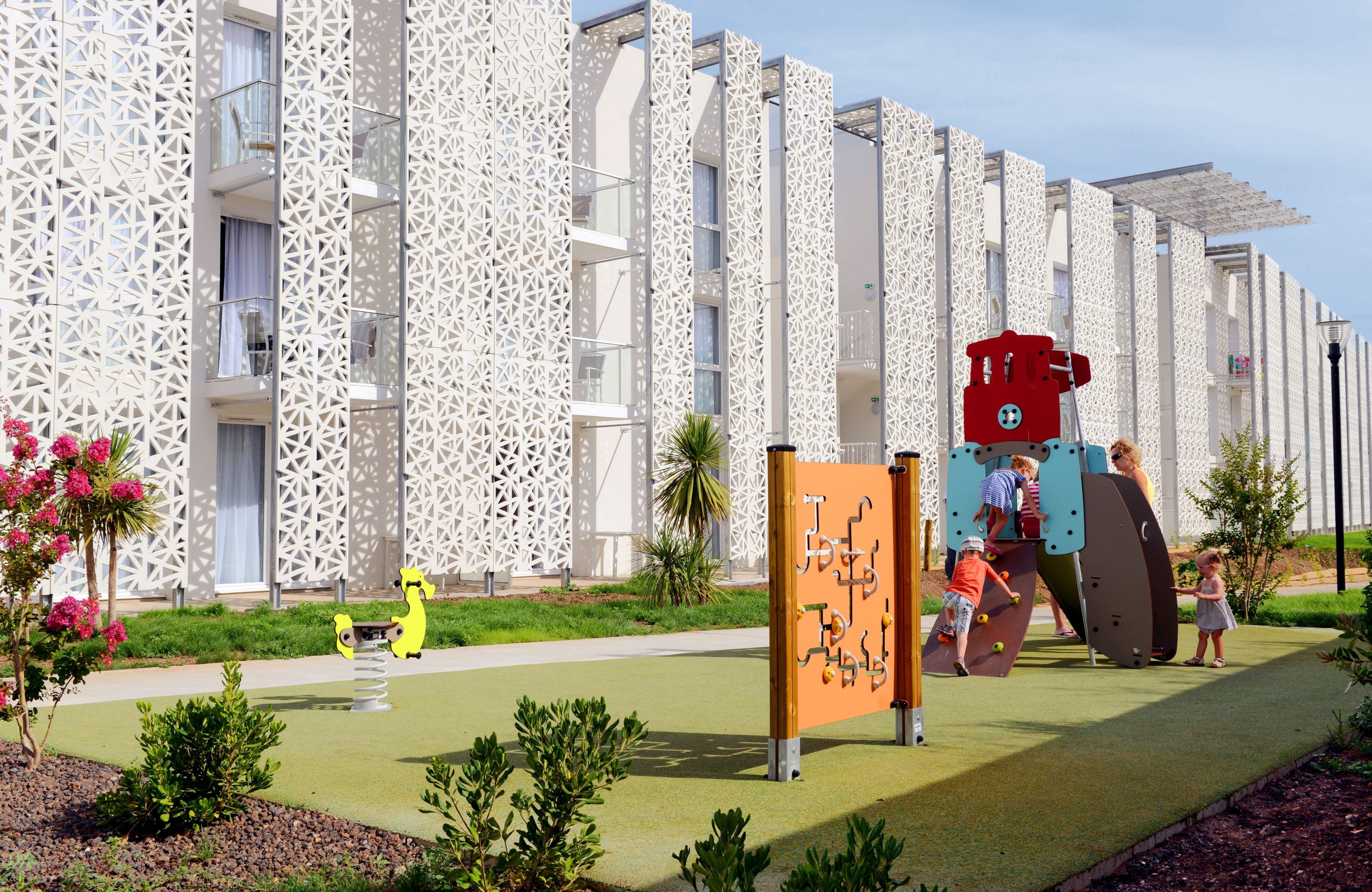A modern building with a unique geometric facade, a children's play area with climbing structures, and a well-maintained garden with small shrubs and flowers.