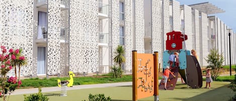 A modern building with a unique geometric facade, a children's play area with climbing structures, and a well-maintained garden with small shrubs and flowers.