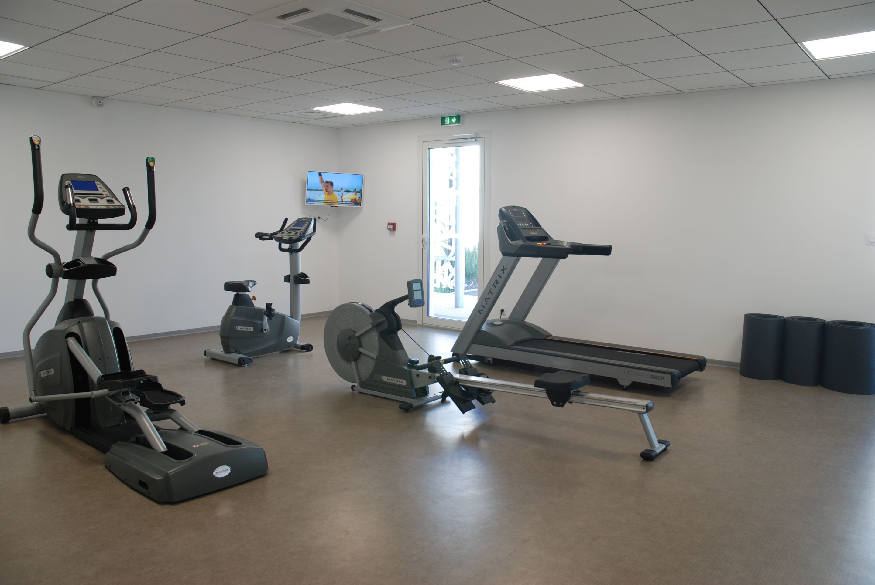 A fitness center with various cardio machines, a television screen, and a door with an exit sign.