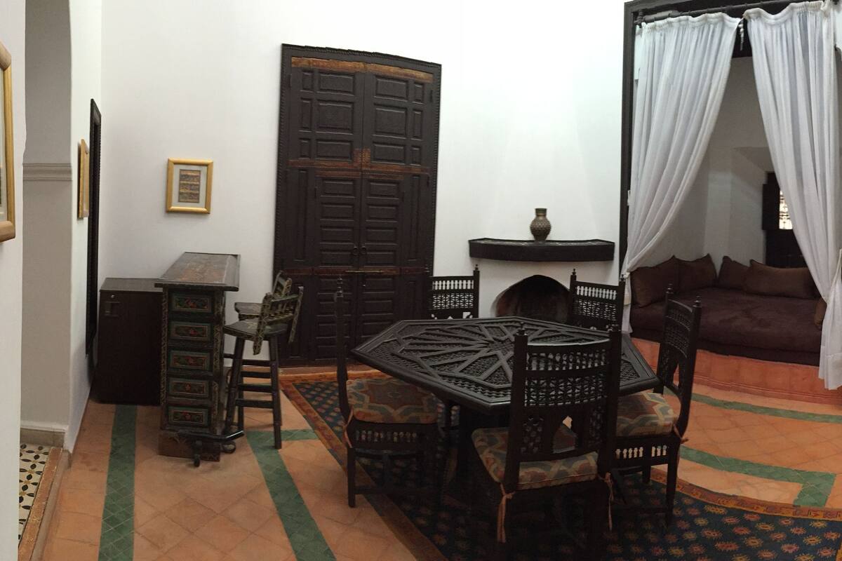 A traditional Moroccan interior with a dining area featuring a low table and chairs, a sofa, and a decorative rug.