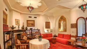 A traditional living room with a red carpet, a round table with a white tablecloth, and a red sofa. There are bookshelves filled with books and decorative items, and a fireplace with a mantel. - Riad Dar El Assafir (Marrakech)