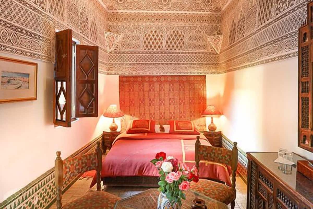 A room with a bed, two bedside tables, a chair, a small table with a vase of flowers, and a decorative ceiling.
