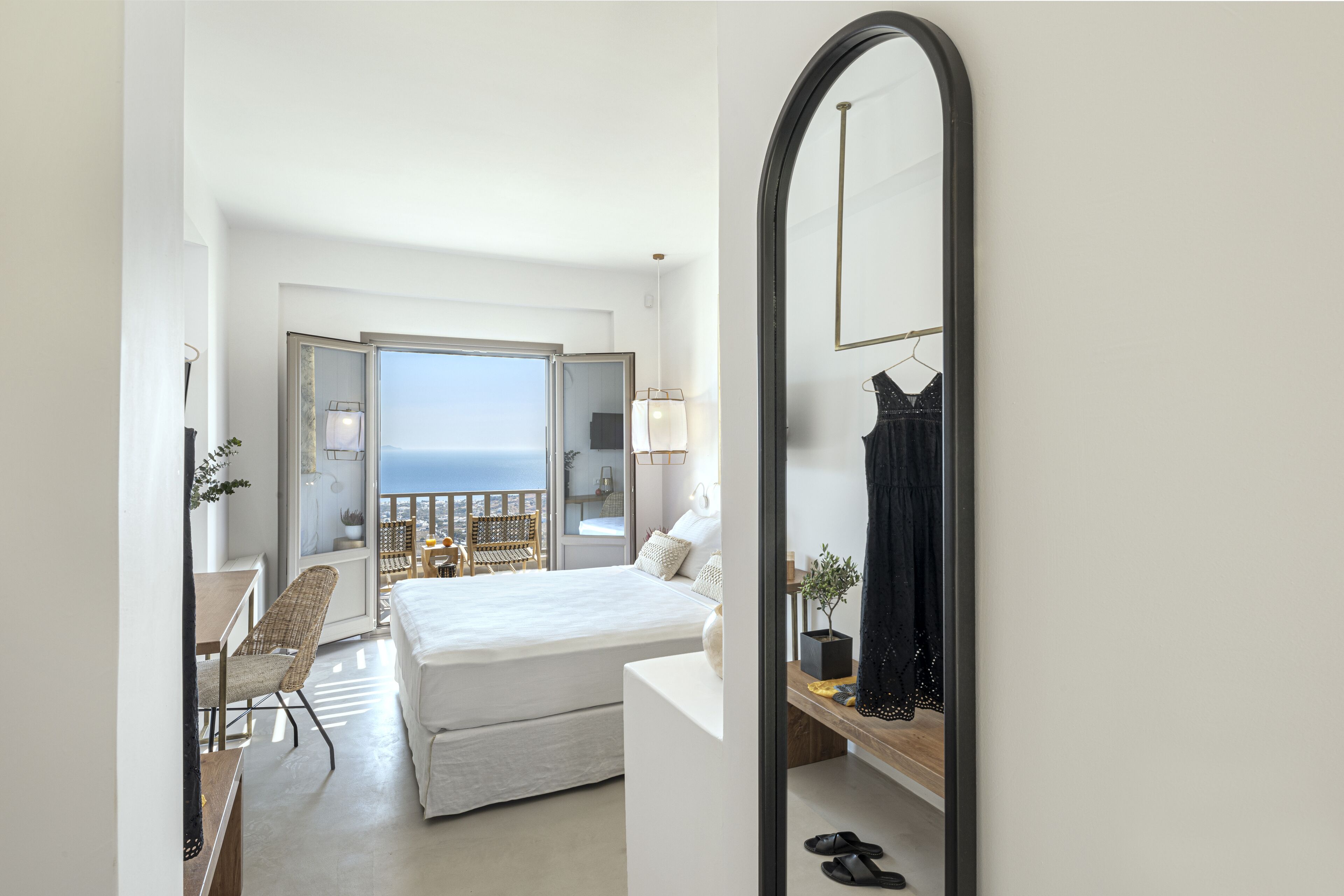 A hotel room with a large bed, a desk, a chair, a mirror, and a view of the sea through a large window.