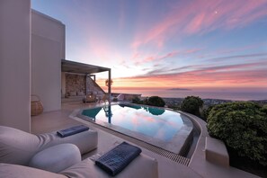 A modern outdoor pool area with a stone wall, a sofa, and a view of the sunset. - Eolia Luxury Villas (Santorini)