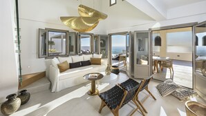 Un amplio espacio de sala con un sofá blanco, un sillón de cuero negro y una mesa de centro redonda. Hay ventanas grandes que ofrecen vistas al océano.