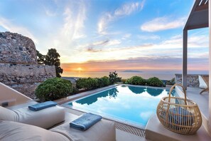 A poolside area with a stone wall, lounge chairs, and a view of the sunset over the sea. - Eolia Luxury Villas (Santorini)
