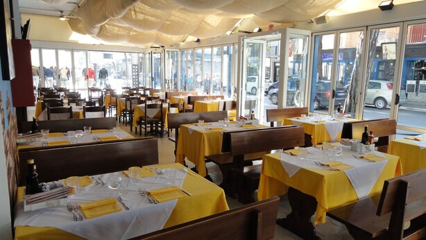 A restaurant with yellow tablecloths, wooden booths, and large windows offering a view of the street. - Hotel Sorrento (Jesolo)