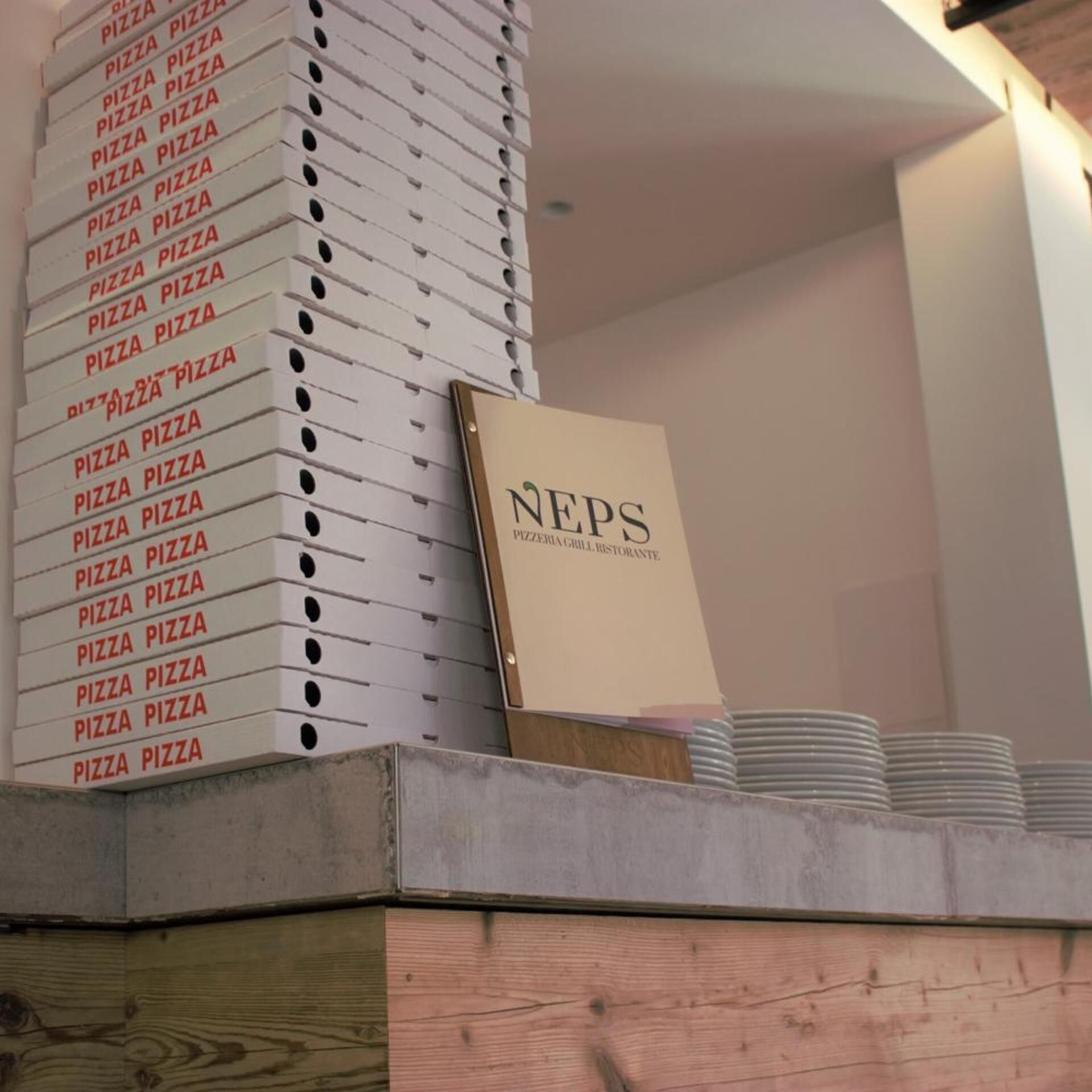 En stabel pizzabokse med teksten 'NEPS', et skilt og tallerkener på en træoverflade.