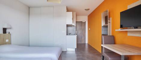 A modern hotel room with a bed, desk, TV, and kitchenette.