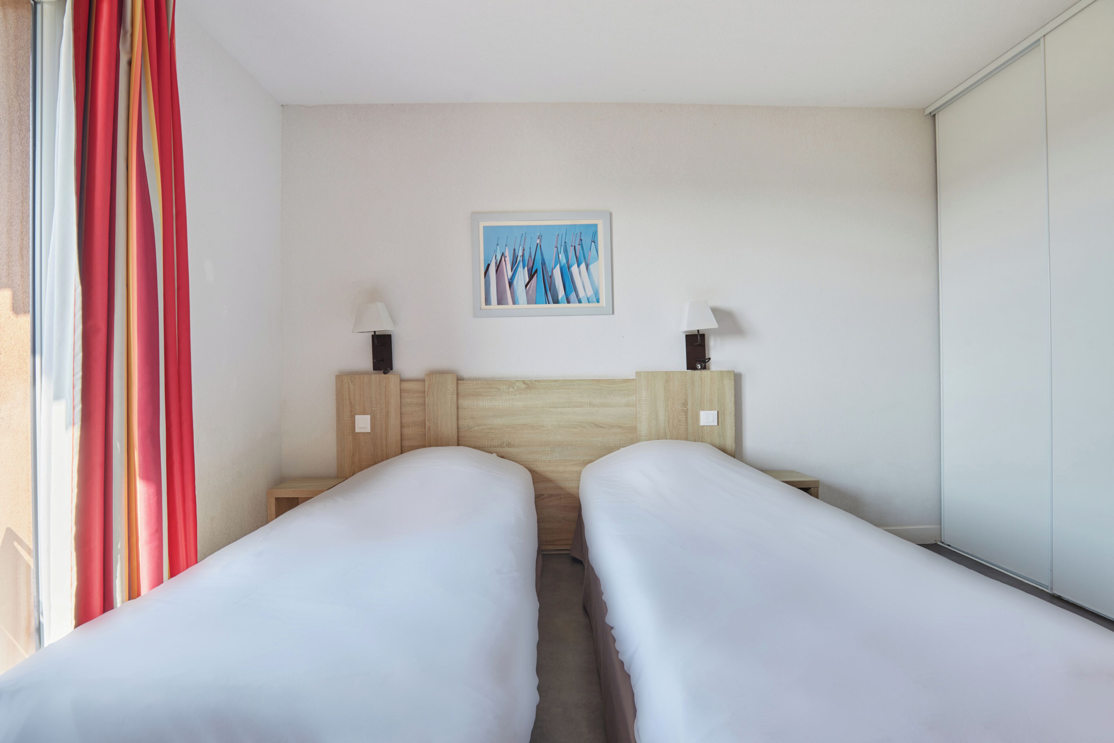 A hotel room with two single beds, a wooden headboard, a painting on the wall, and a window with curtains.