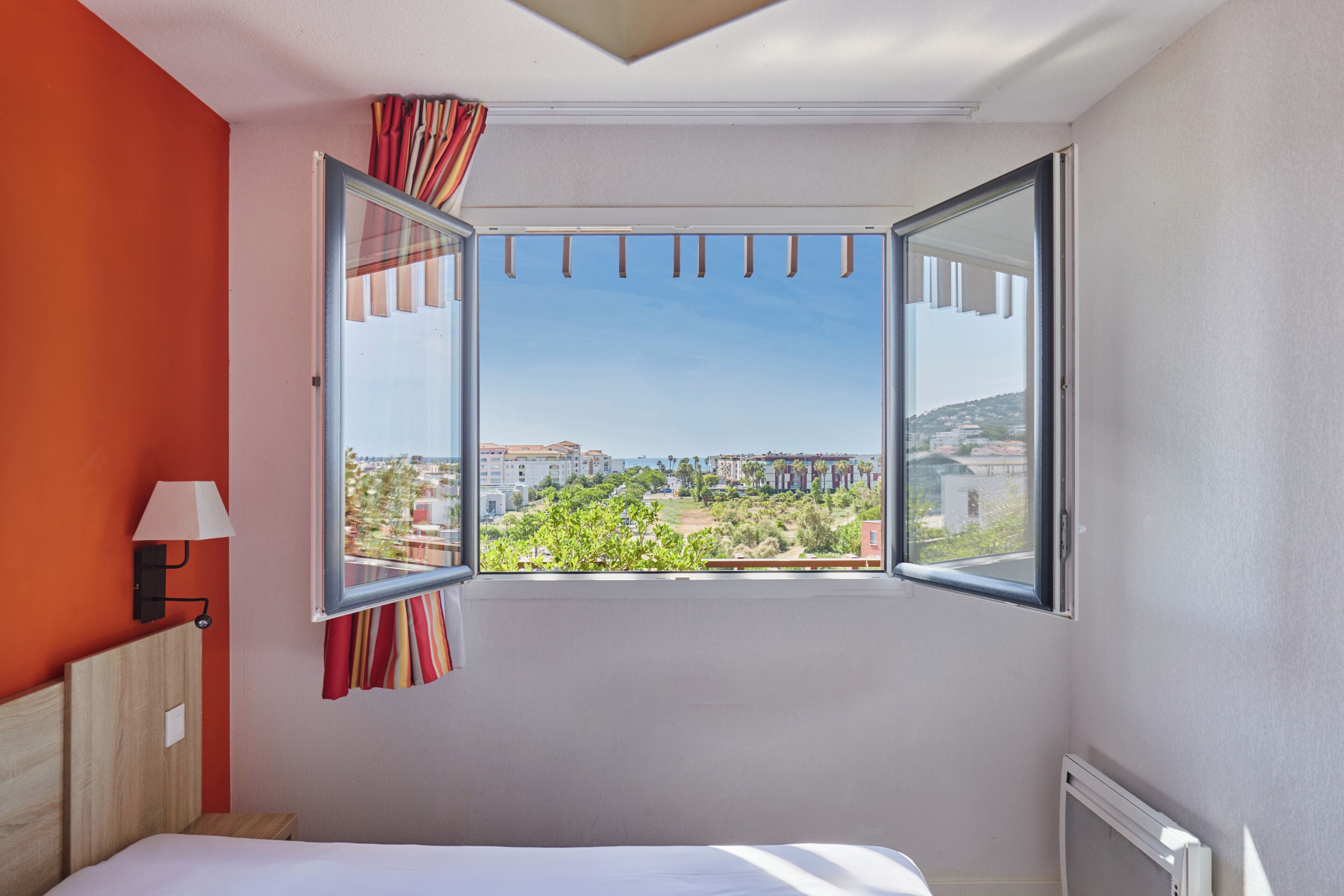 A room with a bed, a window with curtains, a lamp, and a view of buildings and greenery.
