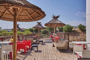A wooden deck with thatched umbrellas, red and white chairs, and a view of greenery and buildings.