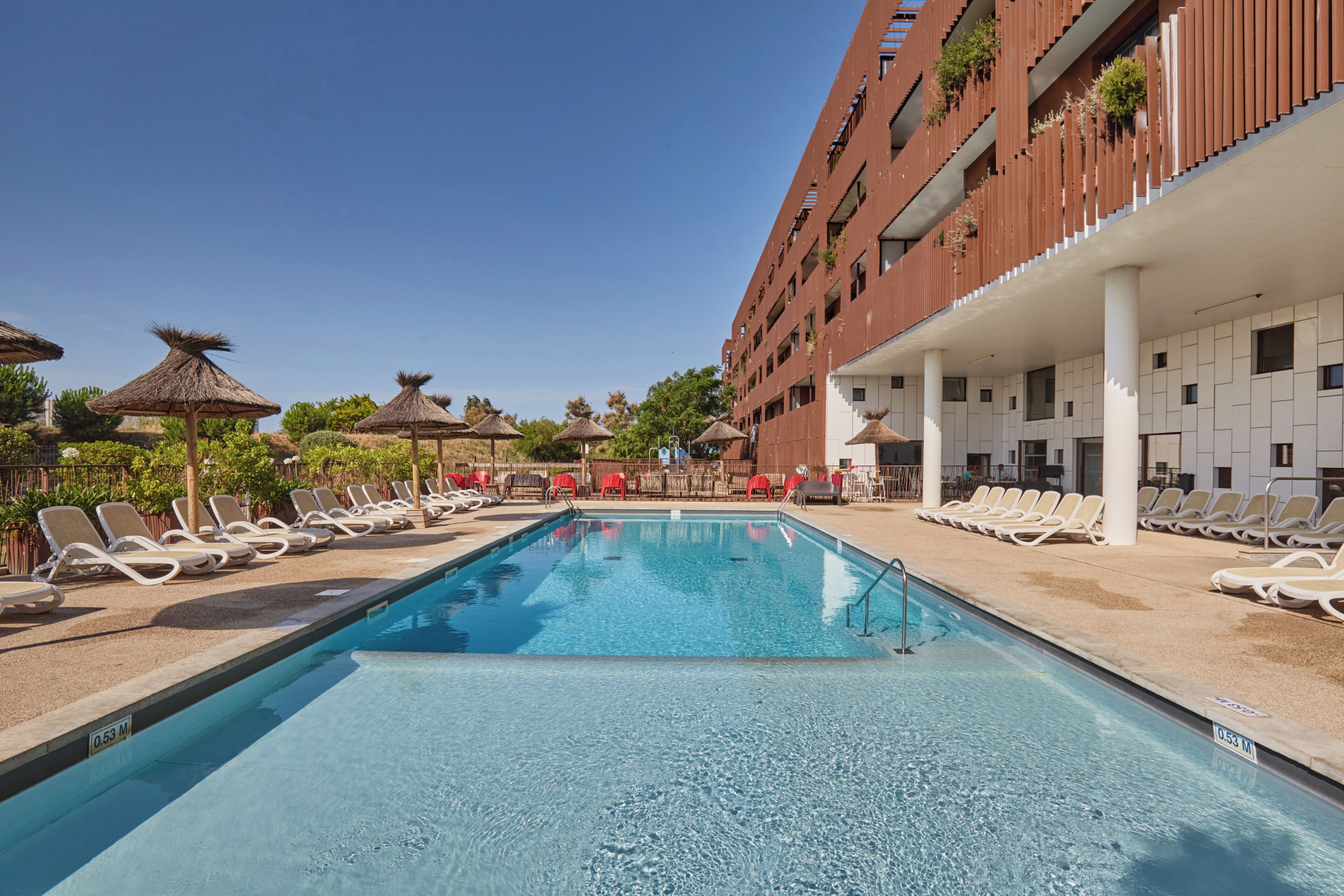 Une piscine entourée de chaises longues et de parasols, elle-même bordée d’un bâtiment doté de balcons, le tout sous un ciel dégagé.