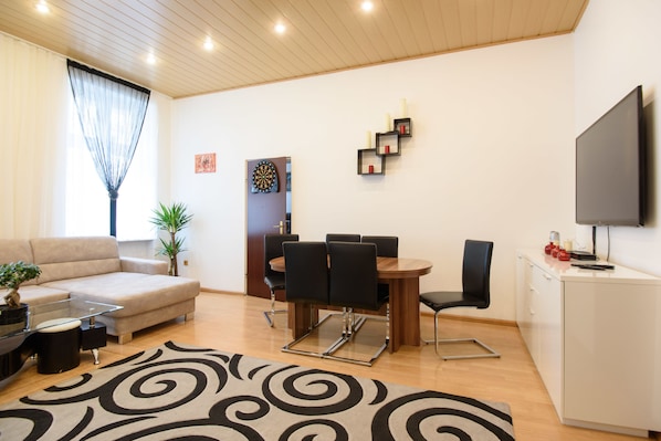 A modern living room with a sofa, coffee table, and dining table with chairs. There is a TV mounted on the wall, a plant, and a decorative wall piece.