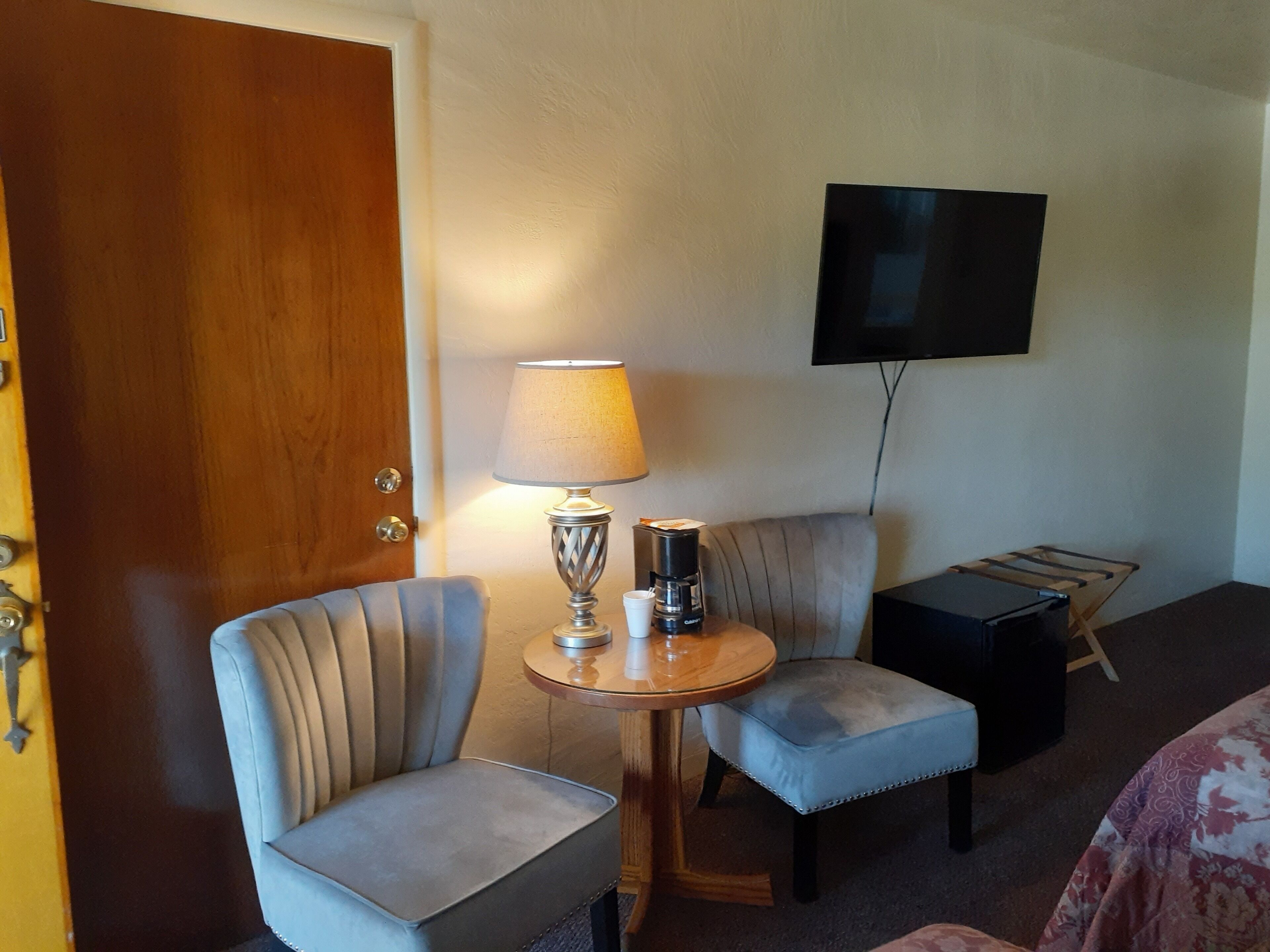 A hotel room with a wooden door, a round table with a lamp, two upholstered chairs, a coffee maker, a flat-screen TV mounted on the wall, and a small wooden side table.