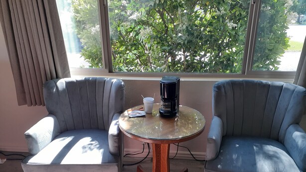 Une pièce avec deux fauteuils gris, une table ronde en verre sur laquelle se trouve une machine à café, et une fenêtre donnant sur des arbres.