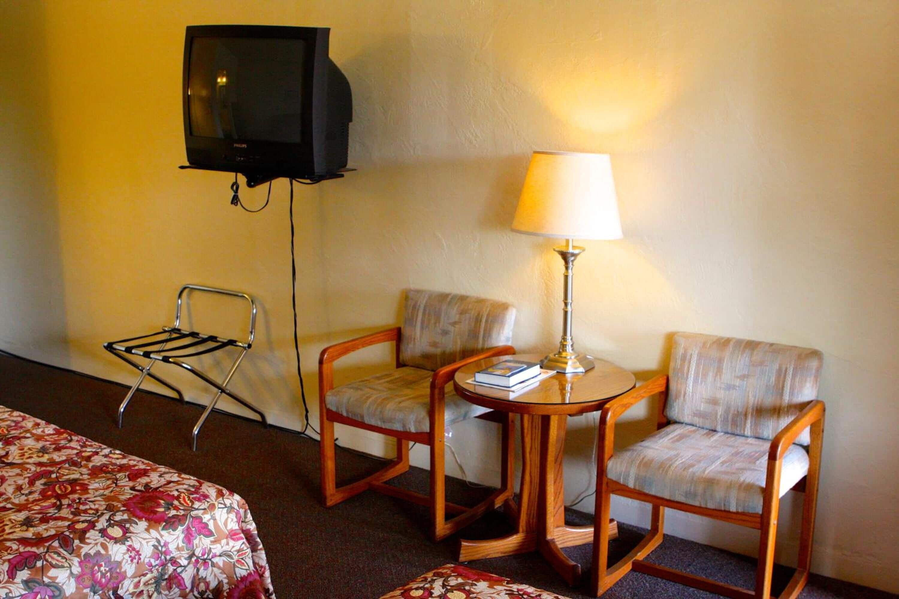 A hotel room with a bed, two armchairs, a small table with a lamp, a TV, and a chair.