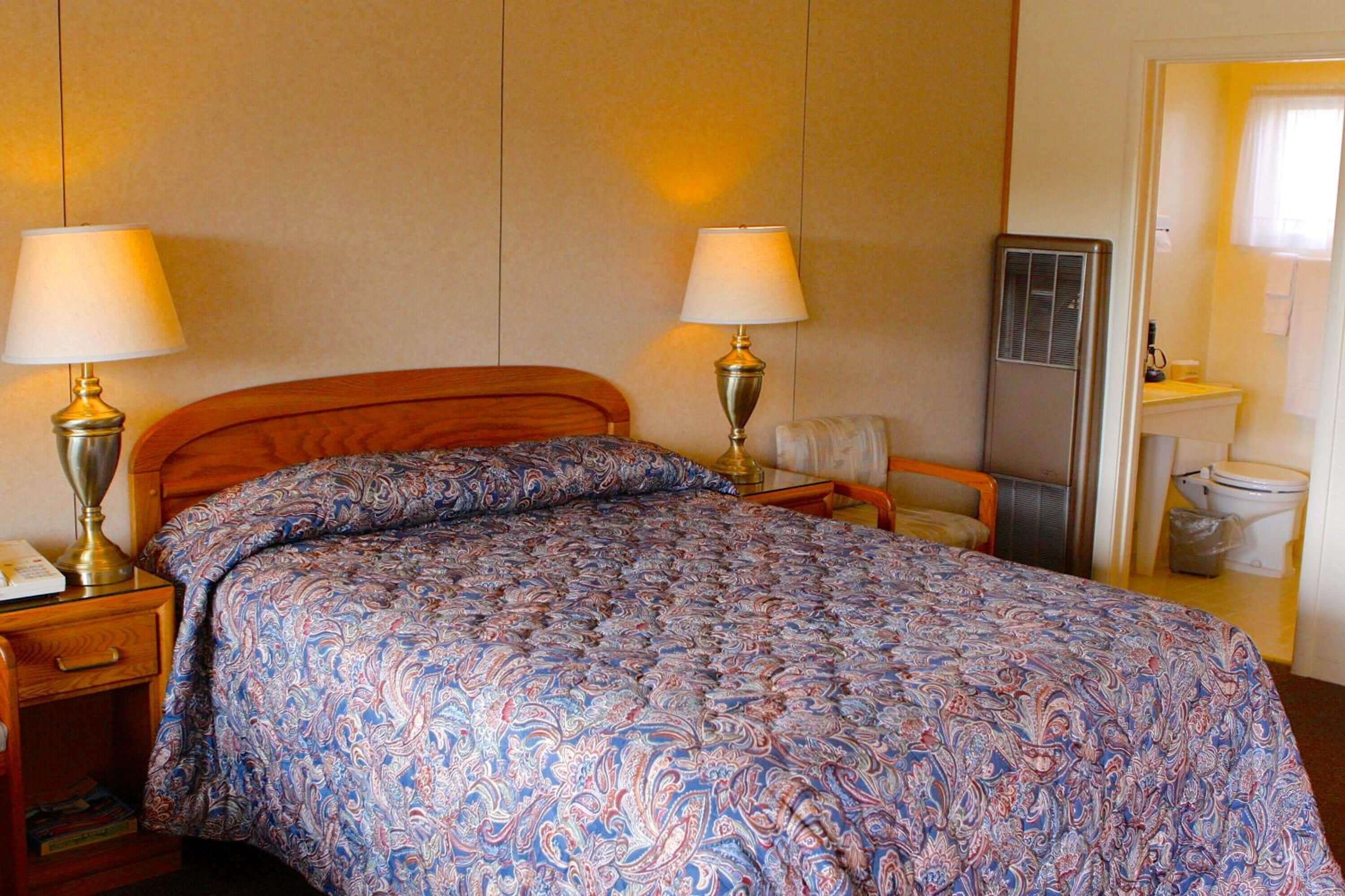 A hotel room with a bed, two bedside lamps, a wooden headboard, a nightstand, a chair, a mirror, and a bathroom with a toilet and sink.