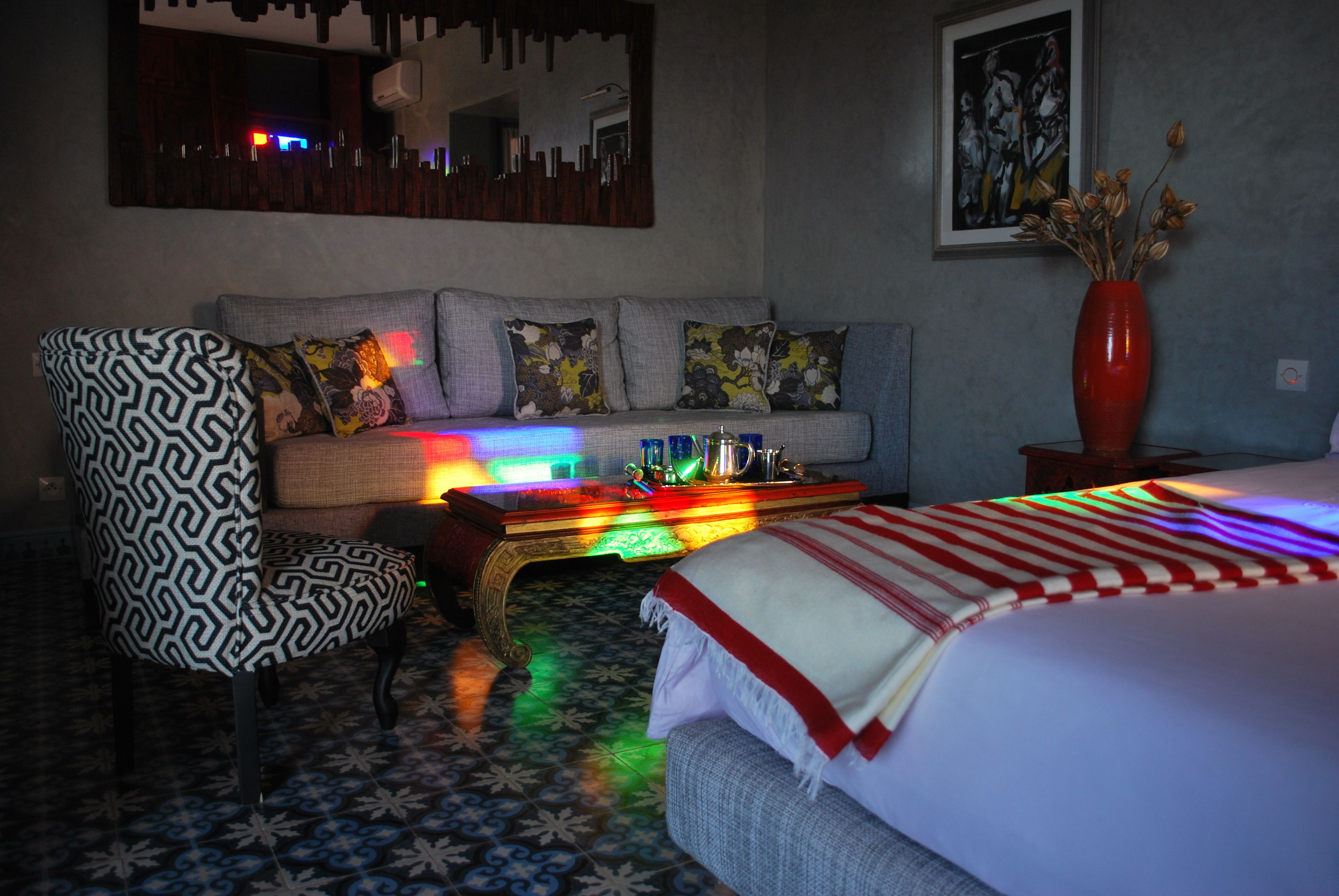 A hotel room with a patterned armchair, a sofa with colorful cushions, a coffee table with a vase, and a bed with a striped blanket.