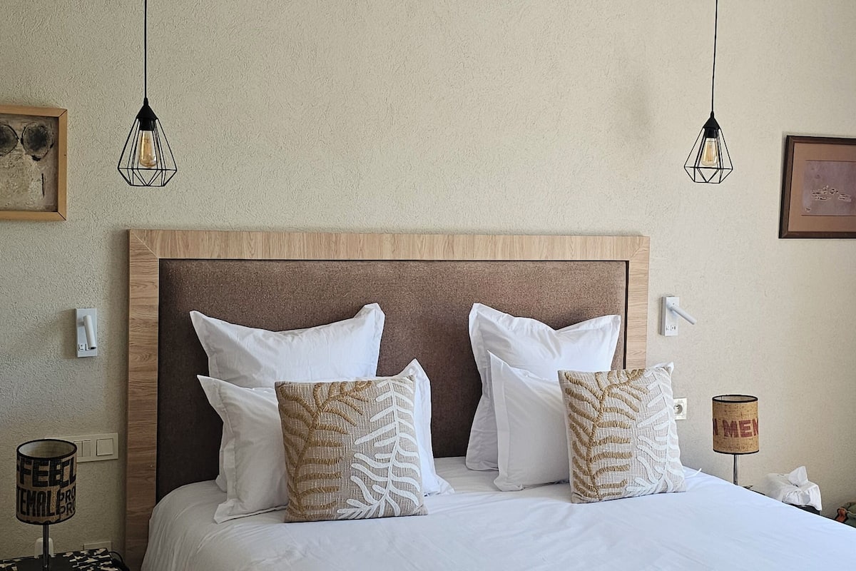 Quarto de hotel com cabeceira de madeira, roupa de cama branca, almofadas estampadas, uma mesa pequena com abajur e obras de arte nas paredes.