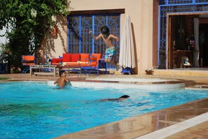 A swimming pool with a child jumping into the water, a lounge area with orange and blue furniture, and a building with blue decorative elements. - Casa Taos (Saâda)