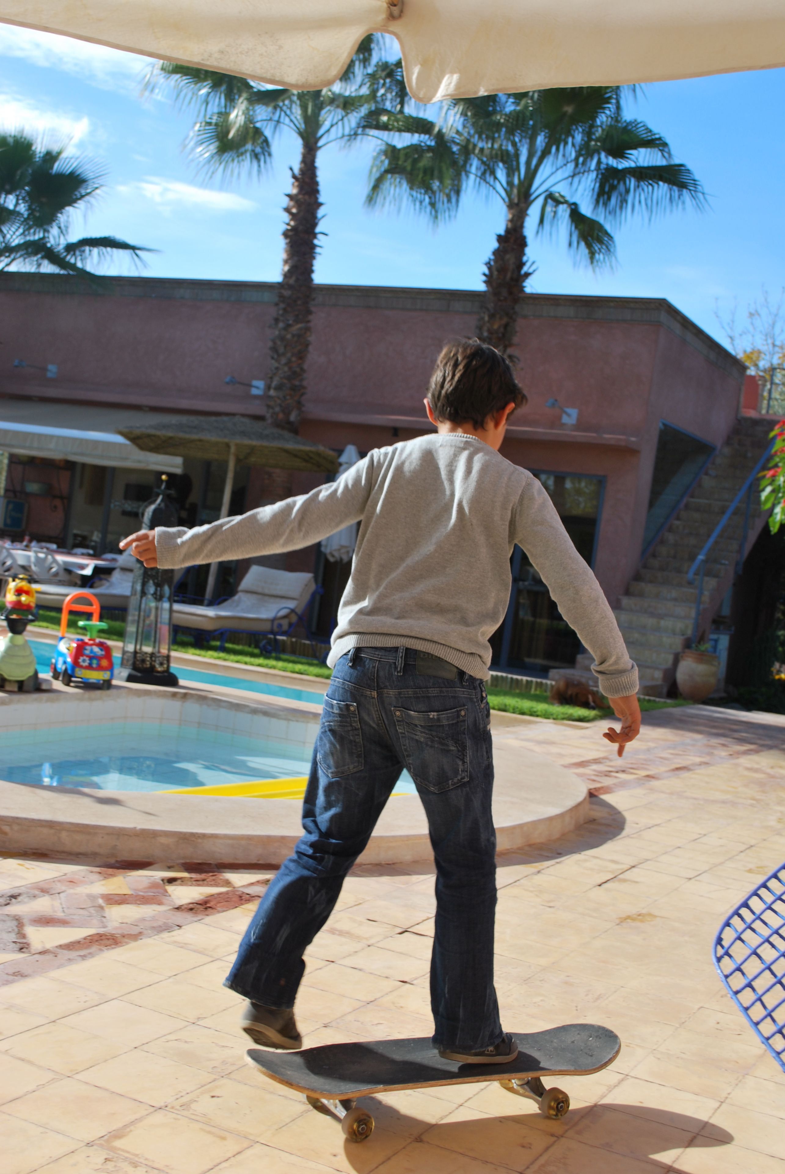 A person skateboarding outdoors near a pool with palm trees and a building in the background.