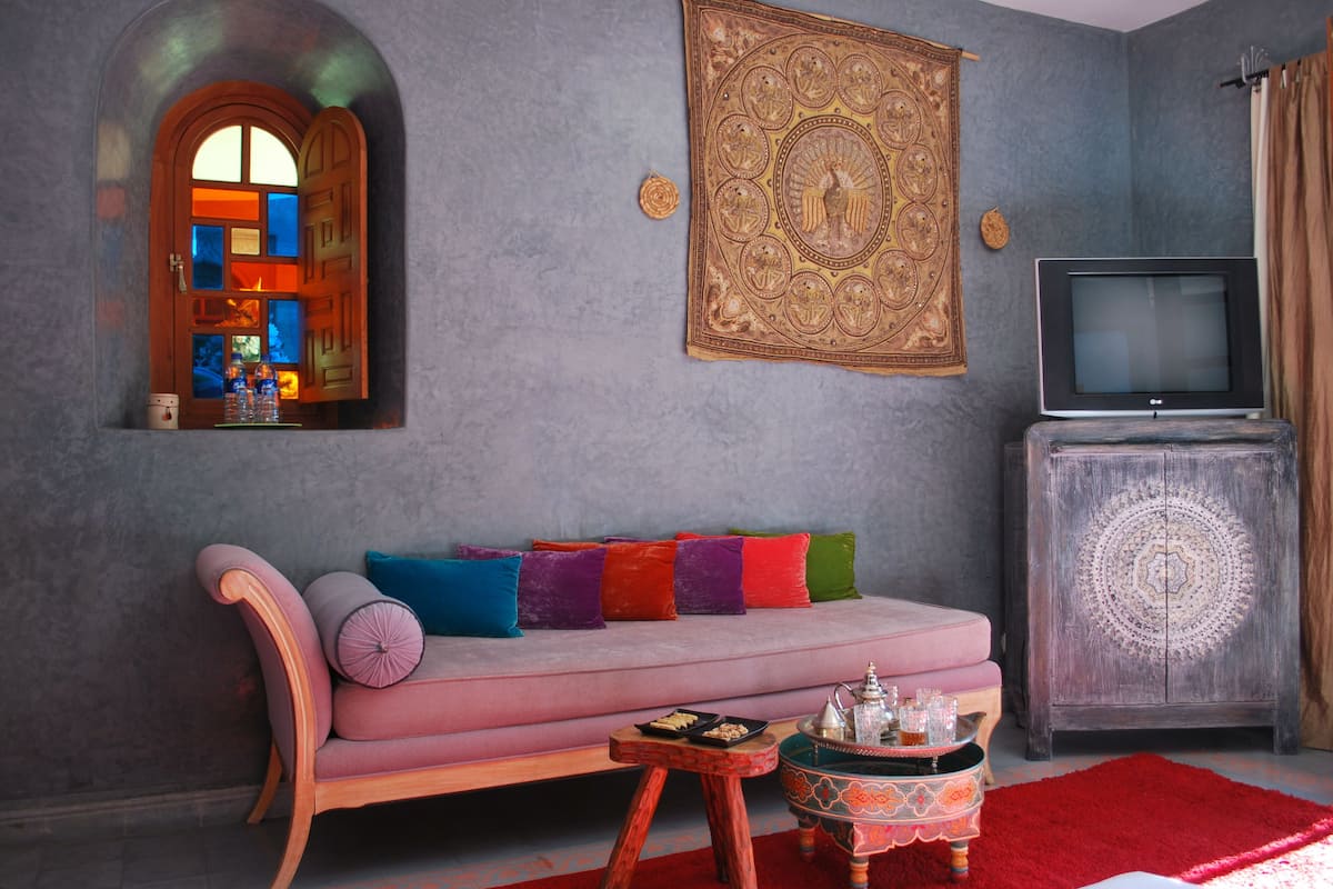 Sala de estar com sofá rosa, almofadas coloridas, uma mesinha com um bule, televisão e uma tapeçaria decorativa.