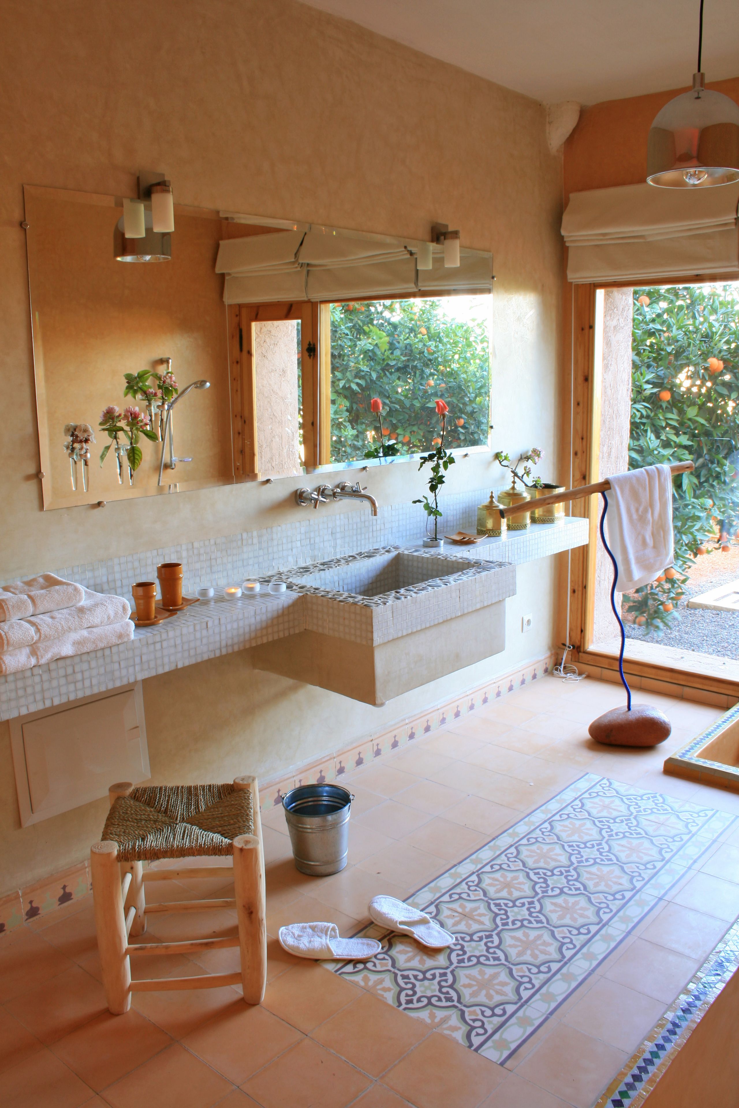 Ein Badezimmer mit einem großen Spiegel, zwei Waschbecken, einer Badewanne, einem hölzernen Hocker, einem Eimer und einem gemusterten Teppich.