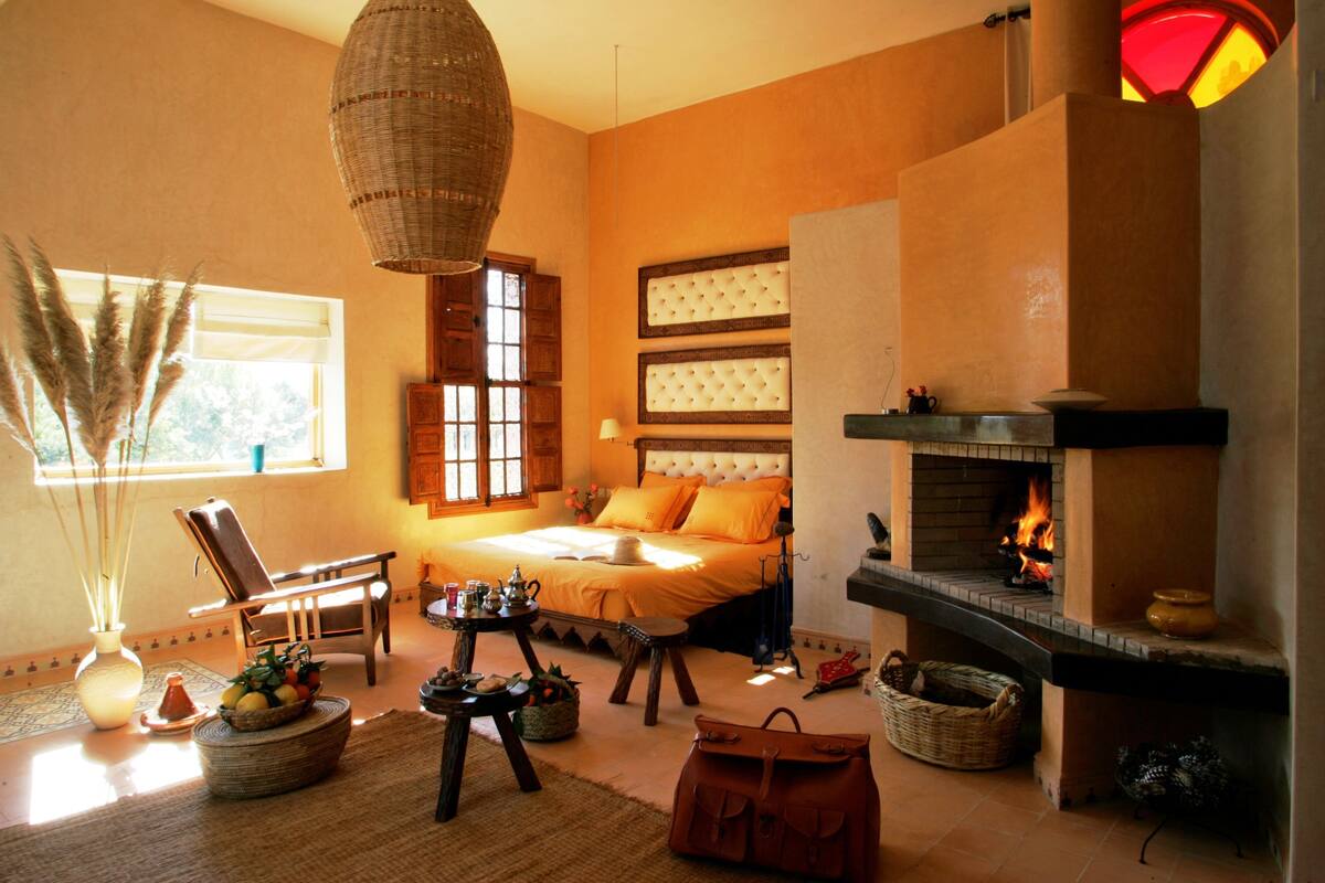 Um quarto aconchegante com lareira, uma cama com roupa de cama laranja, uma cadeira de vime e uma mesa com cesta de frutas.