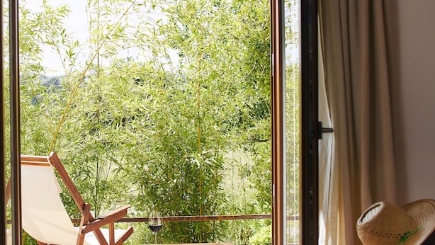 Una habitación con un gran ventanal corredera que da a un jardín, una cama, una silla, una mesa y un sombrero de paja sobre una silla.