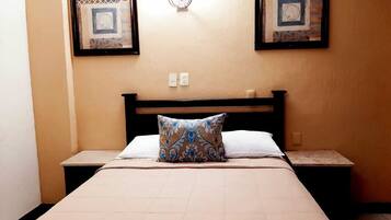 A hotel room with a bed, two bedside tables, a nightstand, a wall-mounted light fixture, and two framed artworks above the bed.