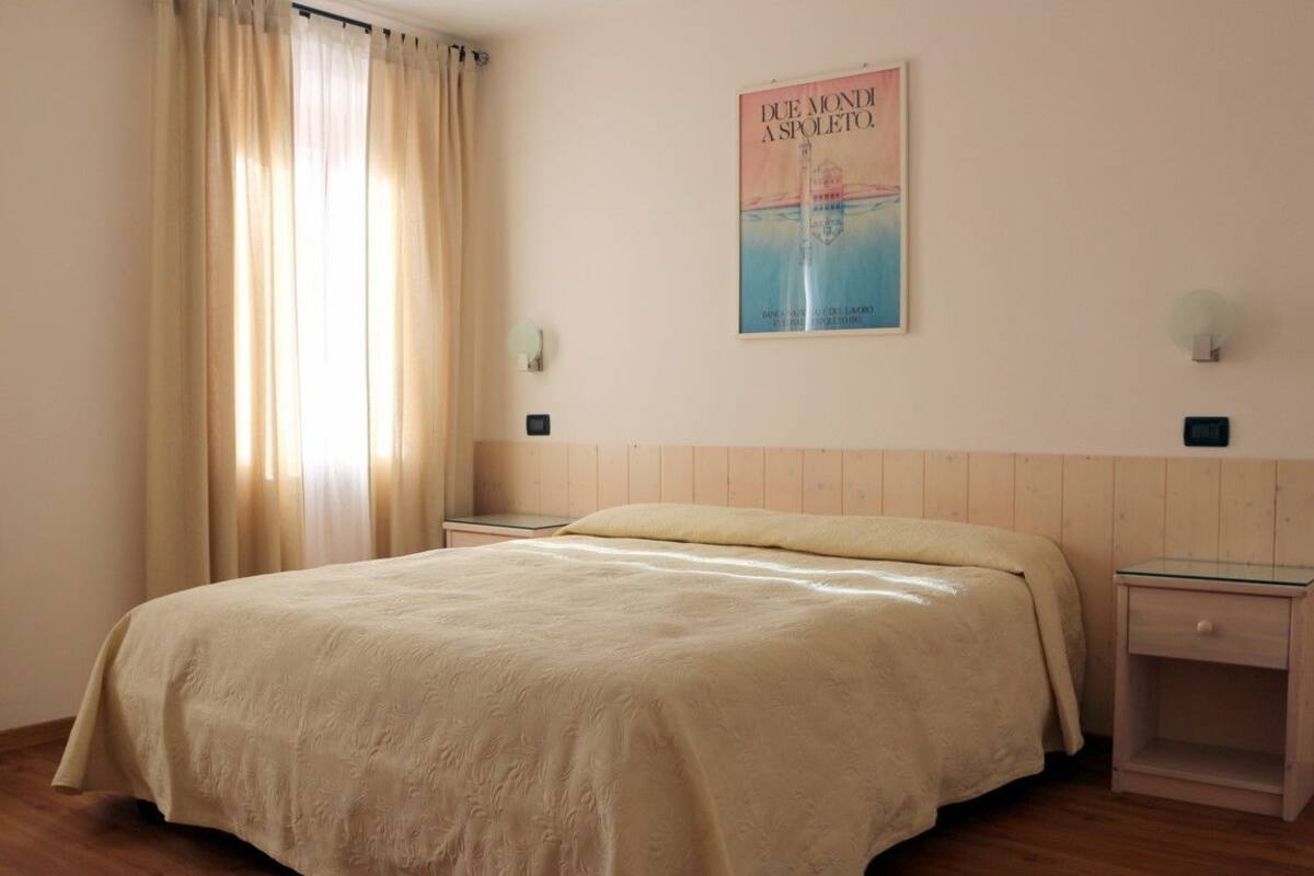Un dormitorio con una cama grande, cabecera de madera, dos mesitas de noche, un cartel en la pared y una ventana con cortinas.