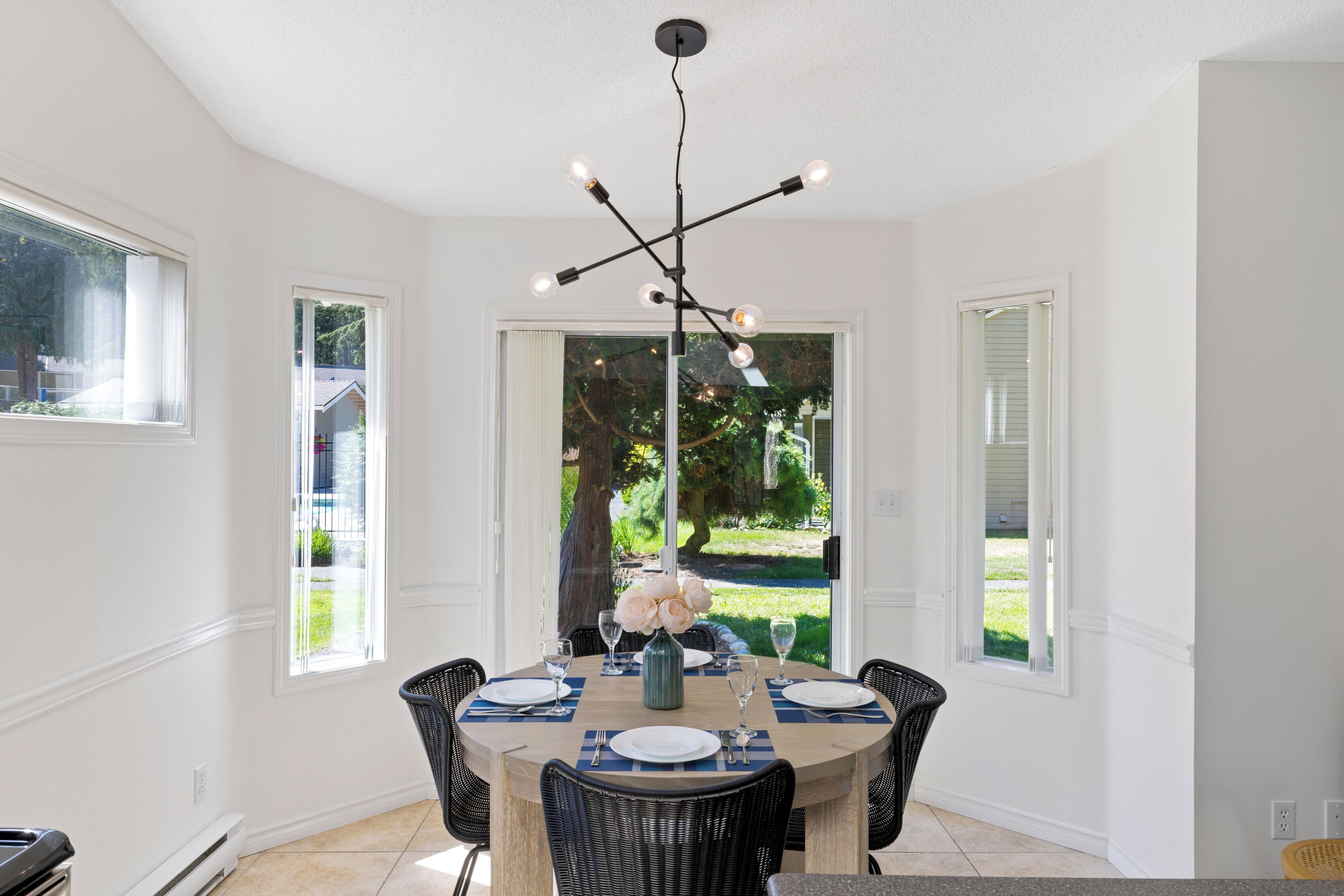 Un coin repas avec une table ronde dressée pour quatre personnes, entourée de chaises en osier, d’un lustre moderne et de grandes fenêtres offrant une vue sur l’extérieur.
