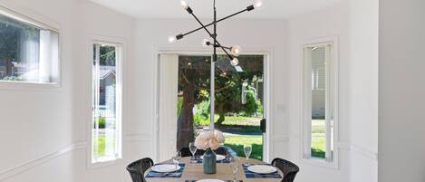 Un coin repas avec une table ronde dressée pour quatre personnes, entourée de chaises en osier, d’un lustre moderne et de grandes fenêtres offrant une vue sur l’extérieur.