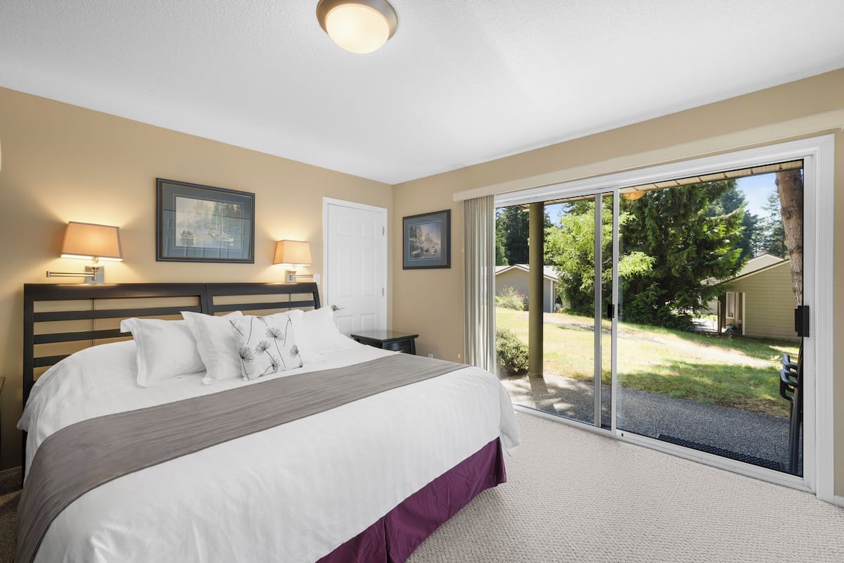 A bedroom with a large bed, two bedside lamps, a door, and a view of a backyard through a sliding glass door.