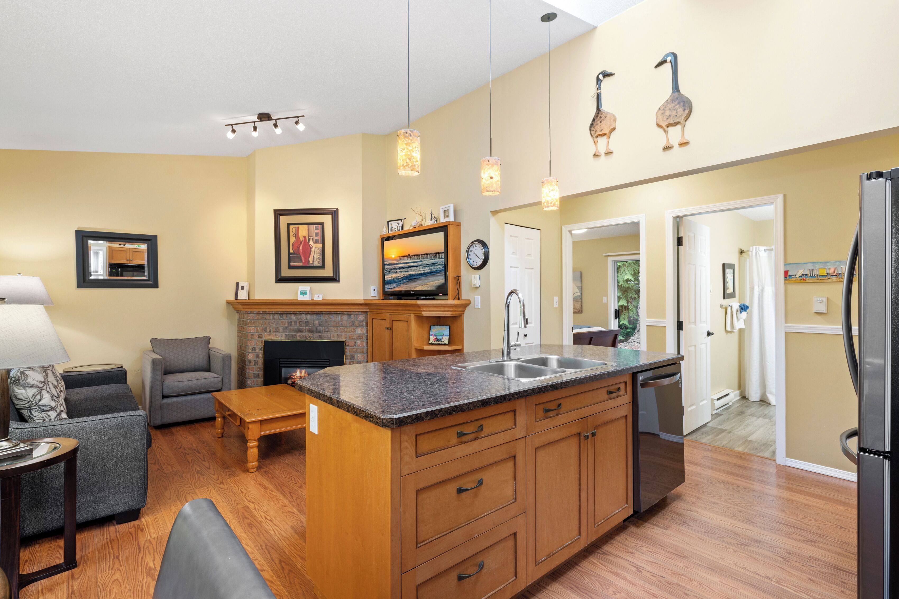A modern kitchen with a central island, wooden cabinets, and pendant lighting. There is a dining area with a table and chairs, and a living room with a sofa and a fireplace.