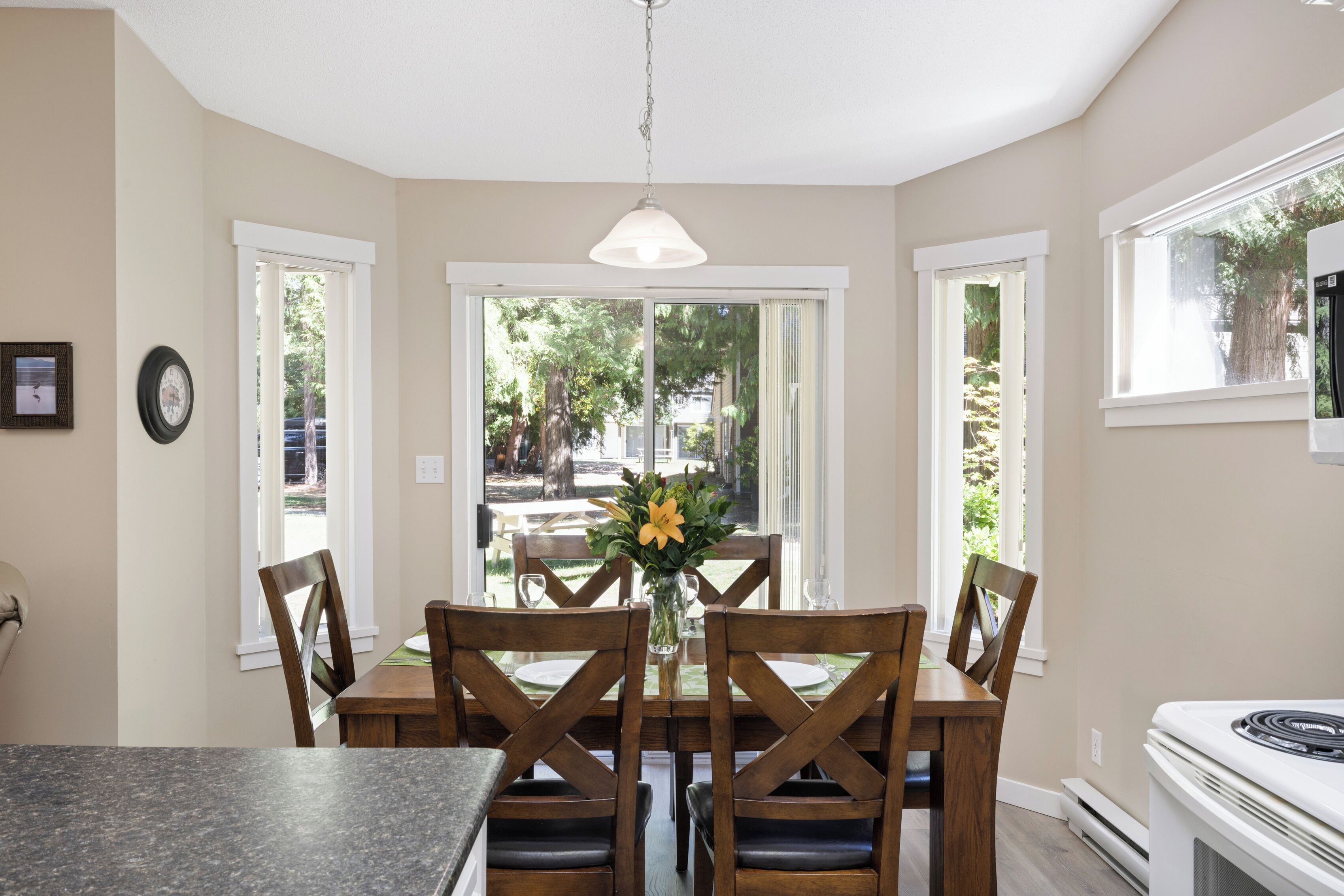 A dining area with a table set for four, wooden chairs, a vase with flowers, and a view of the outdoors through a glass door.