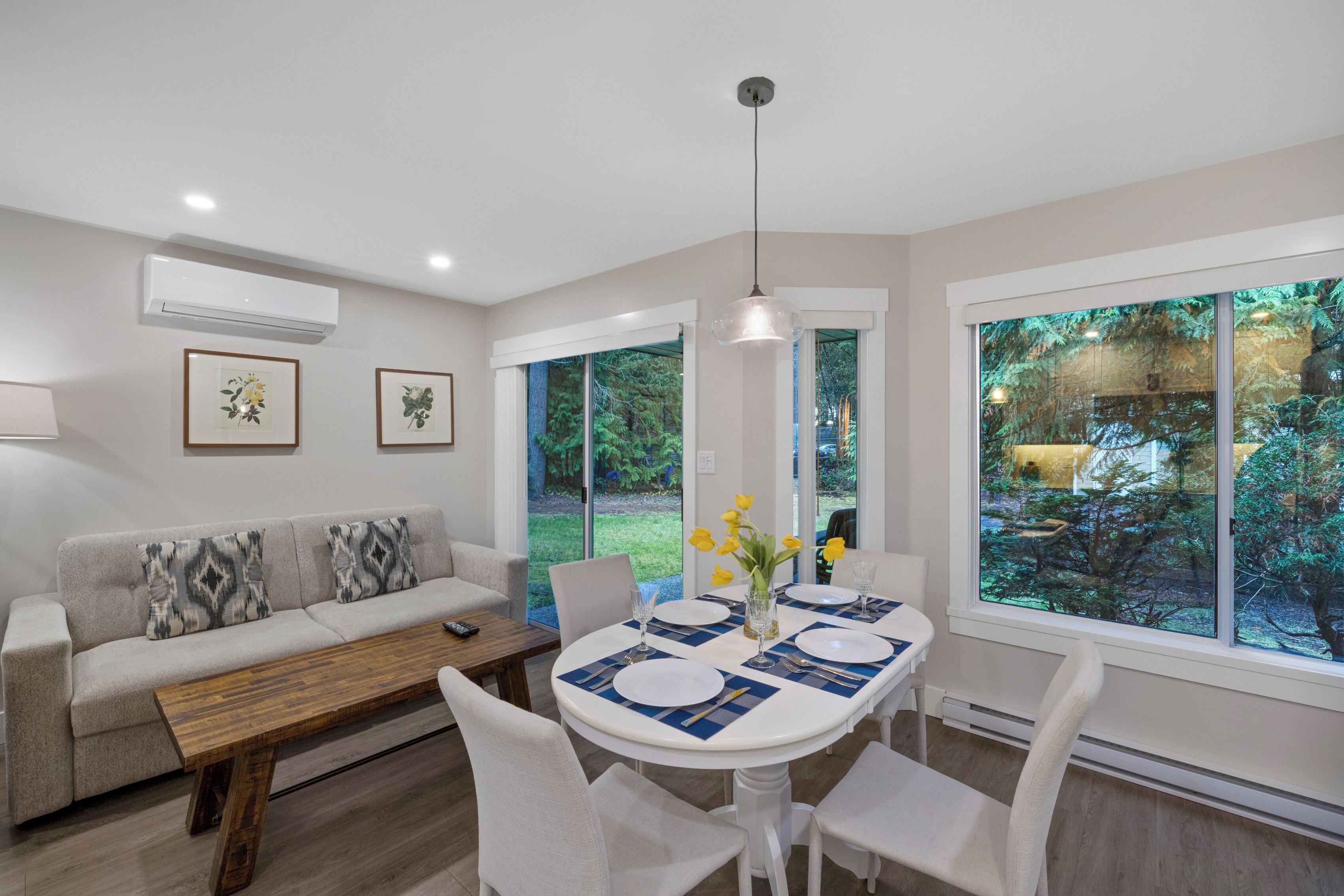 A modern living room with a dining area, featuring a sofa, a wooden coffee table, a dining table set for four, and large windows offering a view of a garden.