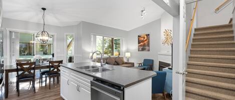Una cocina moderna con una isla central, un comedor con sillas de madera, una escalera y una sala de estar con un sofá y una lámpara.