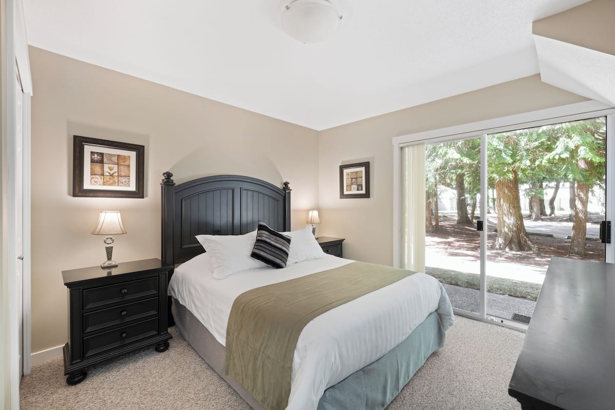 A bedroom with a bed, two bedside tables with lamps, a large window with a view of trees, and a sliding glass door.