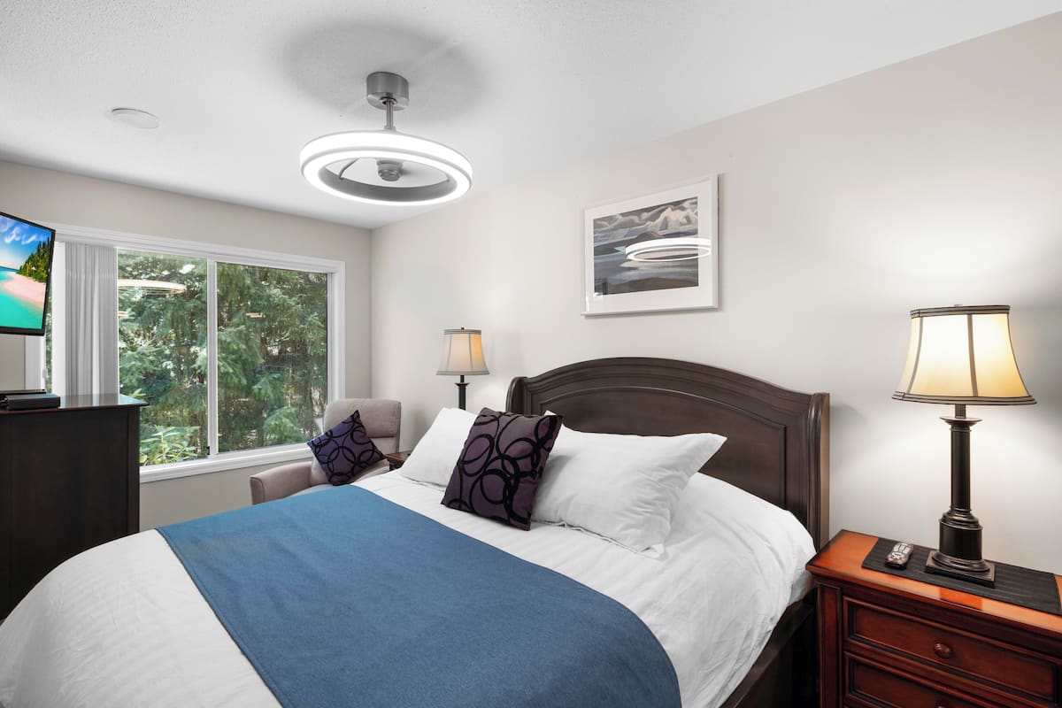 A hotel room with a bed, bedside lamps, a TV, and a window with a view of trees.