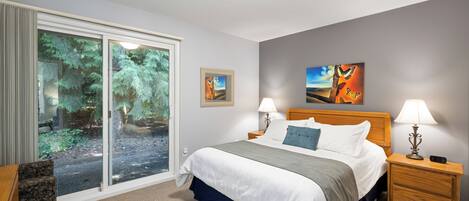 A hotel room with a large bed, two bedside tables with lamps, a chair, a sliding glass door to a garden, and two framed pictures on the wall.