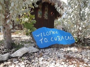 A welcome sign in front of a rustic wooden building with a thatched roof, surrounded by greenery and rocks. - Sea Horizon Apartments (across from Mambo Beach) (Willemstad)