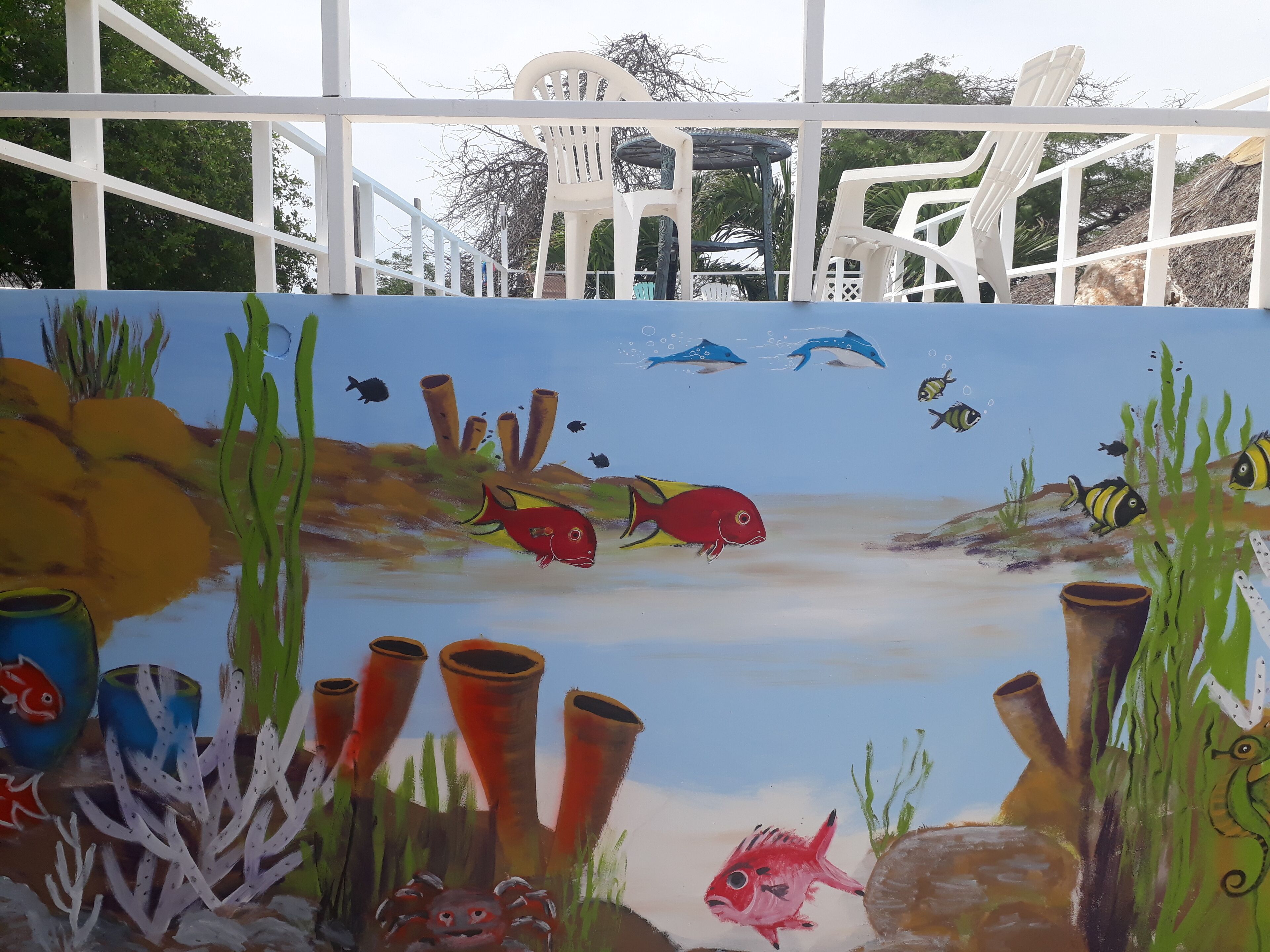 Quarto com uma grande pintura de uma cena subaquática, cadeiras brancas e vista para árvores.