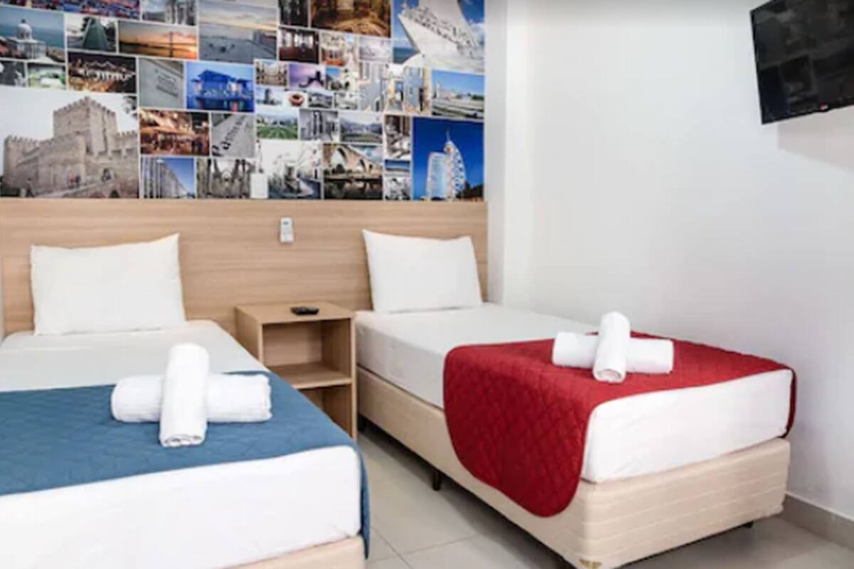 A hotel room with two beds, a wall of travel photos, and a TV mounted on the wall.