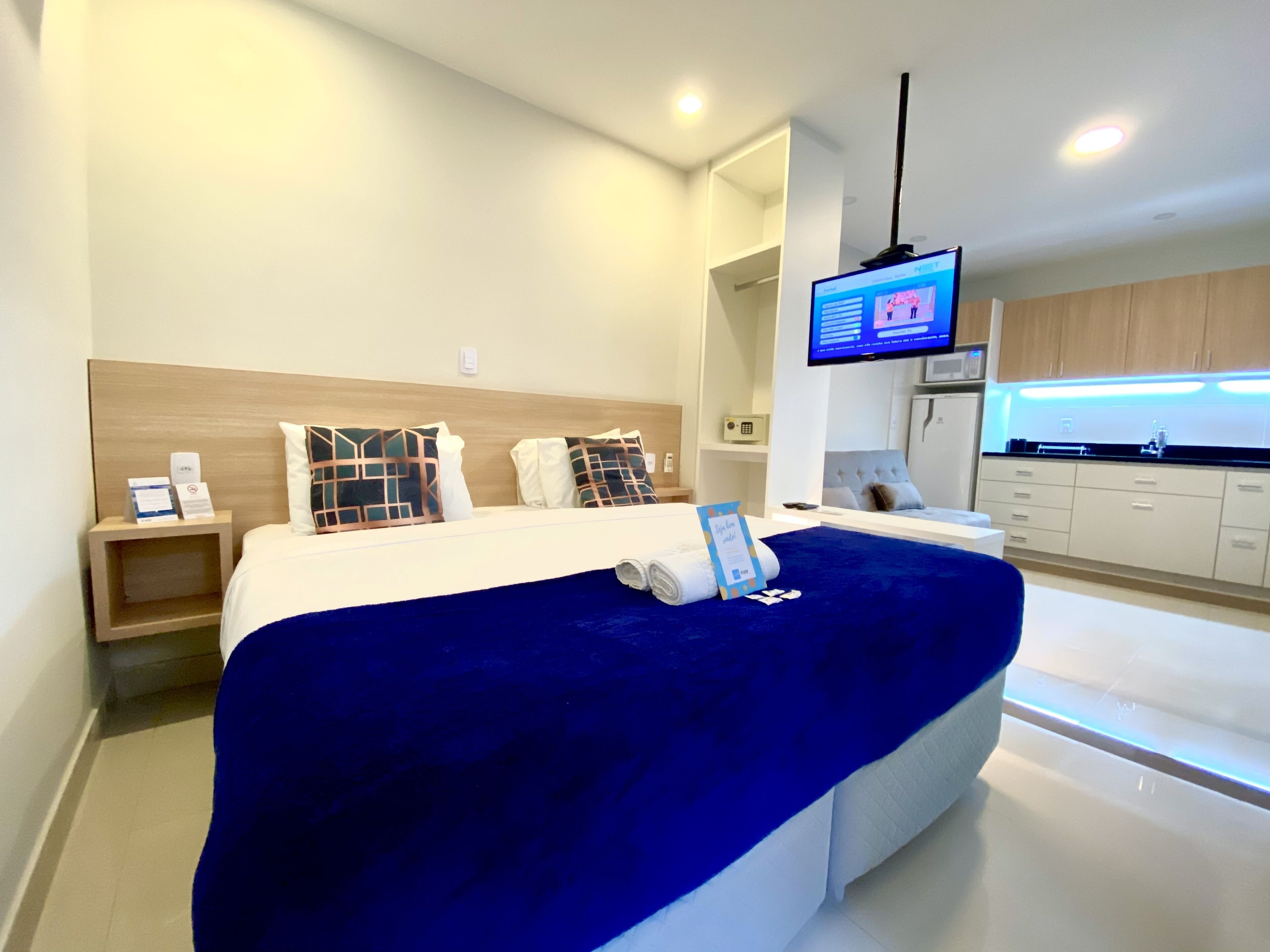 A modern hotel room with a large bed, a TV, a small bedside table, and a view of the kitchen.