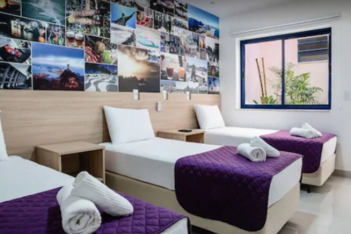 A hotel room with two beds, a large wall of photos, a window with a view of plants, and a bedside table with a lamp.