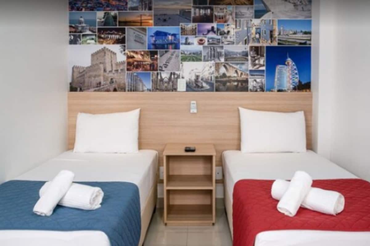 A hotel room with two beds, each with a different colored bedspread, a wooden headboard, and a wall decorated with a collage of travel photos.